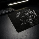Vista oblíqua do Mouse Pad Control Gamer Justice preto com arte ilustrativa em branco e preto de uma figura feminina com uma coroa de espinhos em sua cabeça, segurando uma balança de pratos. Um teclado mecânico preto está visível no canto superior esquerdo da imagem, em uma mesa de madeira escura iluminada por uma luminária de LED.