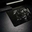 Vista oblíqua do Mouse Pad Control Gamer Justice V3 preto com arte ilustrativa em branco e preto de uma figura feminina com uma coroa de espinhos em sua cabeça, segurando uma balança de pratos. Um teclado mecânico preto está visível no canto superior esquerdo da imagem, em uma mesa de madeira escura iluminada por uma luminária de LED.