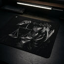 Ângulo para o Mouse Pad Control Gamer Justice posicionado na esquerda da imagem, sua cor é preta com arte ilustrativa em branco e preto de uma figura feminina de cabelos brancos com uma coroa de espinhos, segurando uma balança de pratos. Um teclado mecânico preto está visível no canto superior esquerdo da imagem, em uma mesa de madeira escura iluminada por uma luminária de LED.