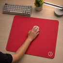 Visão superior de uma mão utilizando um mouse branco sobre o Mouse Pad Gamer Ultraerialace vermelho. O produto está sobre uma mesa de madeira clara, compondo um setup gamer minimalista com teclado claro, luminária de mesa e um pequeno vaso de planta.