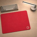 Mouse Pad Gamer Ultraerialace vermelho vazio sobre uma mesa de madeira, capturado em um ângulo levemente inclinado. A iluminação do ambiente realça a textura do tecido e a qualidade das bordas costuradas, com teclado e luminária compondo o cenário ao fundo.