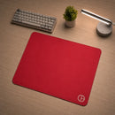 Mouse Pad Gamer Ultraerialace vermelho posicionado em uma mesa de madeira, visto em perspectiva diagonal. A imagem destaca a aderência da base de borracha e o design limpo do mouse pad, acompanhado por um teclado, luminária acesa e planta ao fundo.