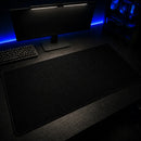 Mouse Pad Grande Gamer Ondas Escuras visto em um ângulo diagonal sobre uma mesa preta. O cenário limpo destaca o produto acompanhado de um teclado mecânico à esquerda, um monitor central e um gabinete de PC com luzes azuis à direita.