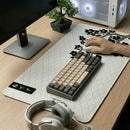 Visão oblíqua de um setup gamer clean com Mouse Pad Grande Gamer Sakura estendido sobre mesa de madeira clara. Sobre o mouse pad, um teclado mecânico compacto e um mouse branco sendo utilizado. Ao fundo, um gabinete de PC branco com iluminação RGB, um monitor e um pequeno bonsai decorativo.