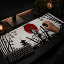 Visão isométrica de um setup de mesa de computador de madeira preta texturizada, com foco no Mouse Pad Grande Gamer Samurai estendido com arte vibrante de estilo sumi-e japonês. A estampa apresenta um samurai em armadura completa, um grande sol vermelho no centro, uma pagode, katanas cruzadas e bordas de bambu em preto e branco. Sobre o mouse pad, há um teclado mecânico preto compacto com uma tecla esc vermelha e um mouse preto sem fio segurado por uma mão humana.