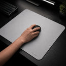 Uma foto em close em ângulo lateral e frontal do Mouse Pad Pk Control Gamer branco sobre a mesa preta, com o logotipo "PK CONTROL I" visível no canto inferior direito e as bordas costuradas brancas bem definidas. O mouse pad se estende sobre a mesa com o teclado mecânico cinza e o vaso de planta desfocados ao fundo, enfatizando a textura da superfície de tecido e o design minimalista.