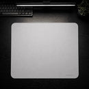 Uma visão aérea centralizada do Mouse Pad Pk Control Gamer branco ocupando a maior parte da cena sobre a mesa preta. Uma luminária linear acesa está acima, o teclado cinza no canto superior esquerdo e o vaso de planta no canto superior direito. O mouse pad retangular branco exibe claramente suas bordas costuradas e o logotipo "PK CONTROL I" no canto inferior direito.
