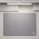 Vista superior do Mouse Pad Speed Gamer Galaxy Gem na cor cinza com textura suave e logo branca no canto inferior direito do produto. O mouse pad repousa sobre uma mesa clara em um setup minimalista, posicionado abaixo de um teclado mecânico branco e uma barra de luz.