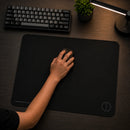 Mão de um jogador utilizando um mouse sobre o mouse pad speed gamer Ultradash preto em uma mesa de madeira escura. O ambiente adequado para jogos inclui um teclado, uma planta e uma luminária.