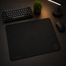 Mouse pad speed gamer Ultradash preto disposto em um setup limpo sobre uma mesa de madeira escurecida. O produto está no centro, acompanhado de perto por um mouse, teclado mecânico, pequena planta e luminária acesa.