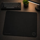 Mouse pad retangular gamer Ultradash preto em uma perspectiva frontal na mesa. O cenário mantém o foco no acabamento suave do produto, com o teclado, planta e luminária dispostos ao redor no fundo escuro.