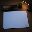 Mouse Pad Speed Ultradash Blue em perspectiva angular sobre uma mesa de madeira. Foco no design limpo e na textura do tecido azul com bordas costuradas. O ambiente mantém o pad como principal destaque, com teclado e luminária desfocados ao fundo.